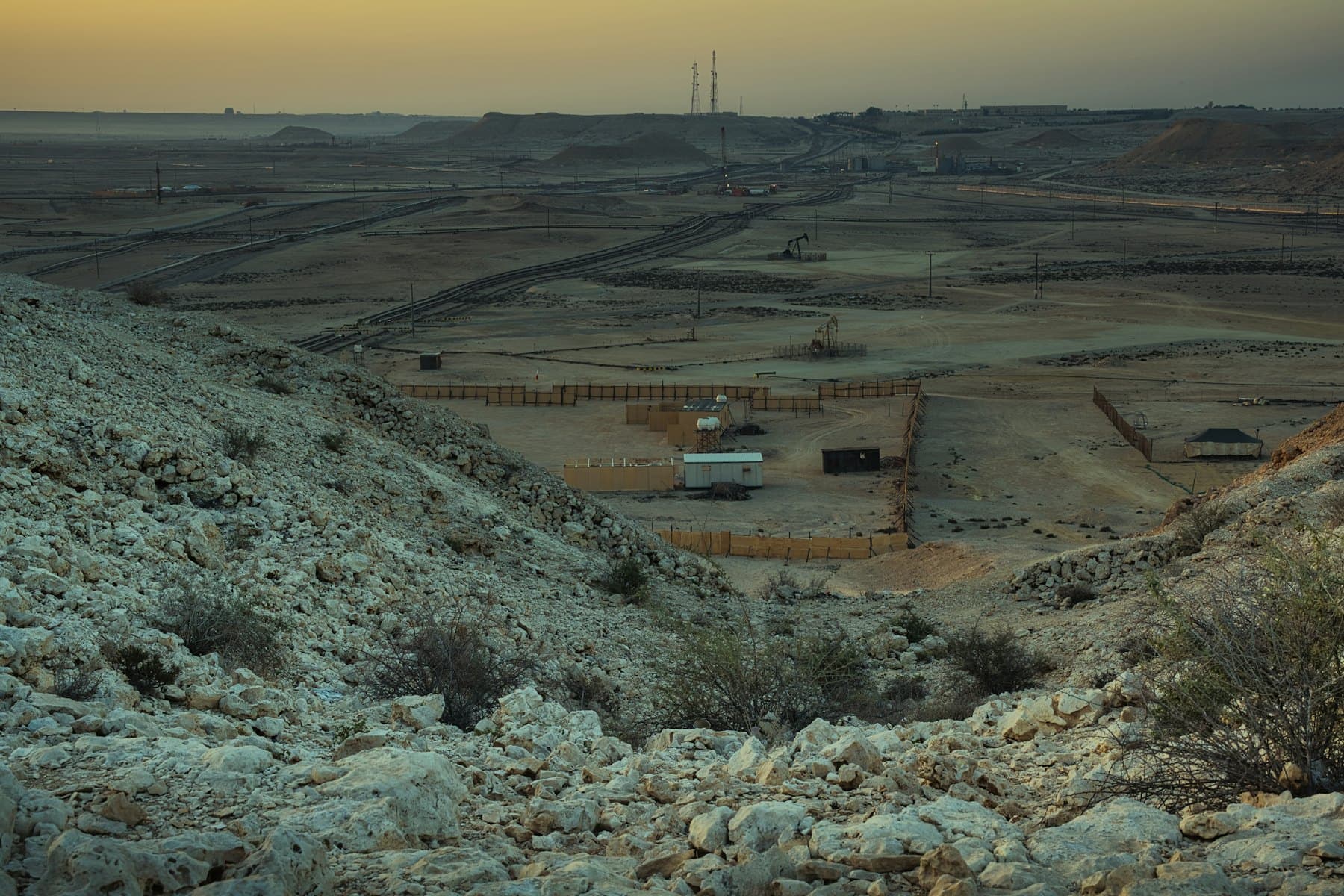 A desert work site with tracks and structures seen from a rocky ridge at dusk